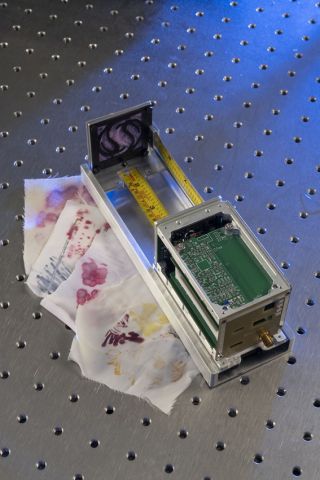 Examples of the fabric painted with bacterial dyes sit beside Spinning Around's SpinnyONE satellite which will carry them into space in February 2026.